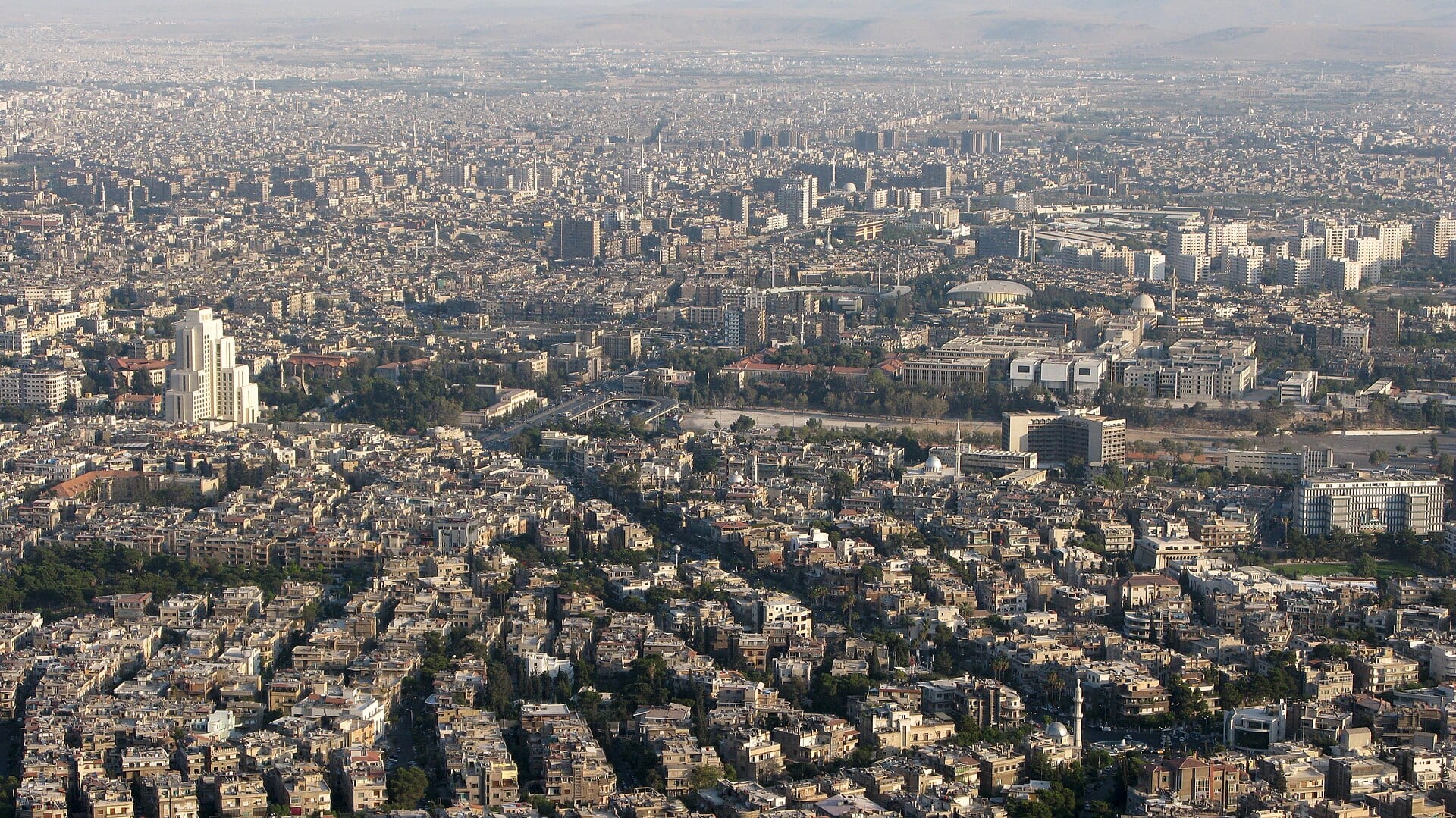 Damascus seen wide from above, the city spreading to the foot of the mountain.