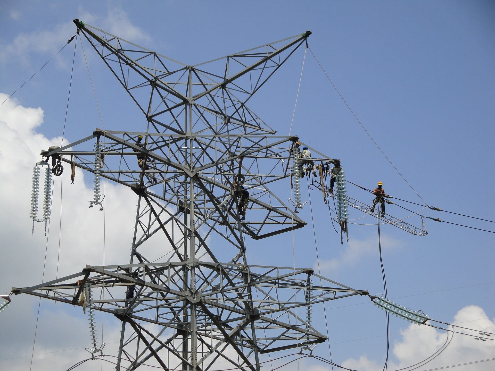 Linesmen in helmets and harnesses working along the crossarms of a transmission tower, insulator strings hanging beside them.