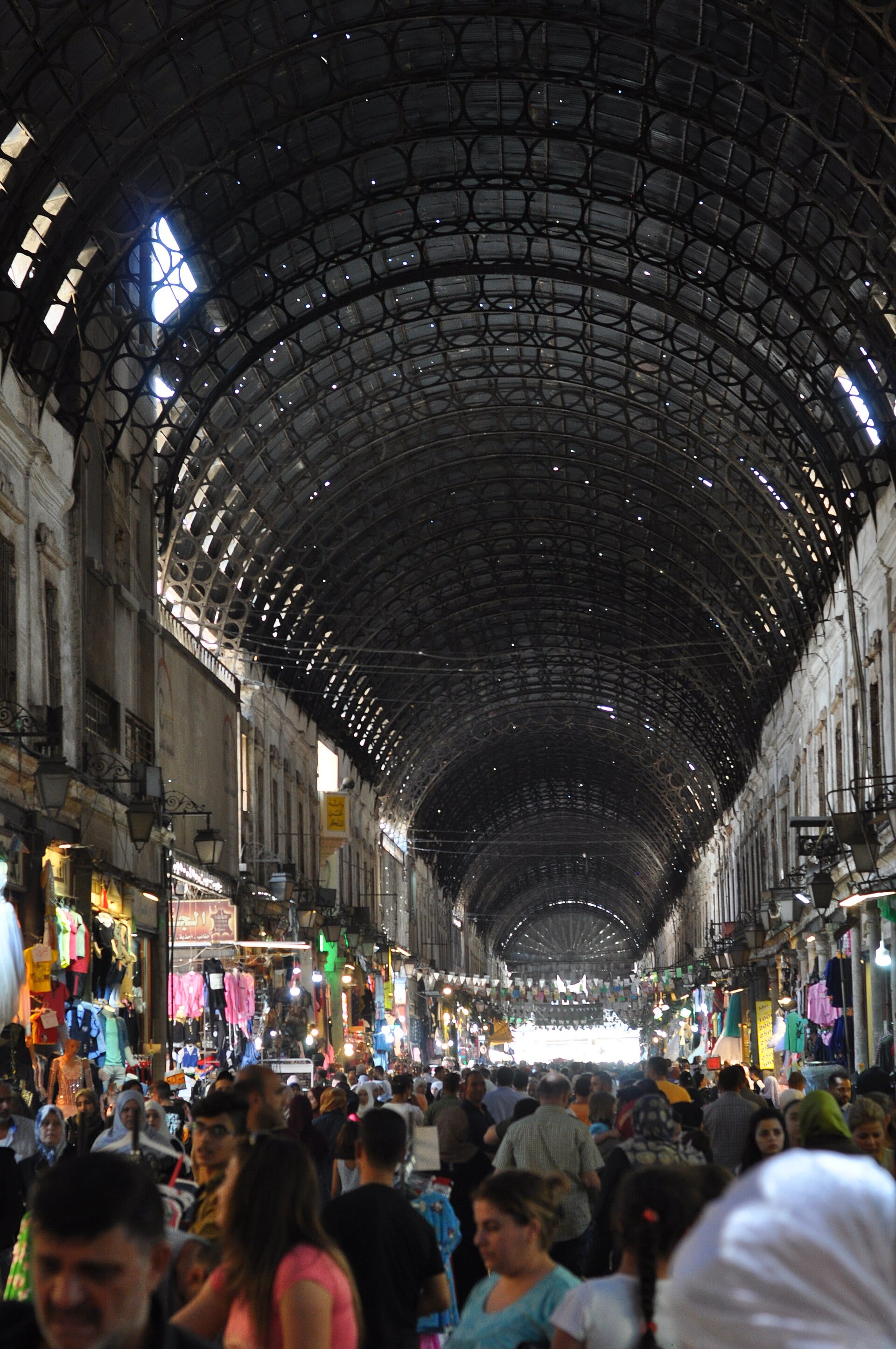 Crowds walking beneath the pierced iron roof of Souq al-Hamidiyah, points of daylight falling through it.
