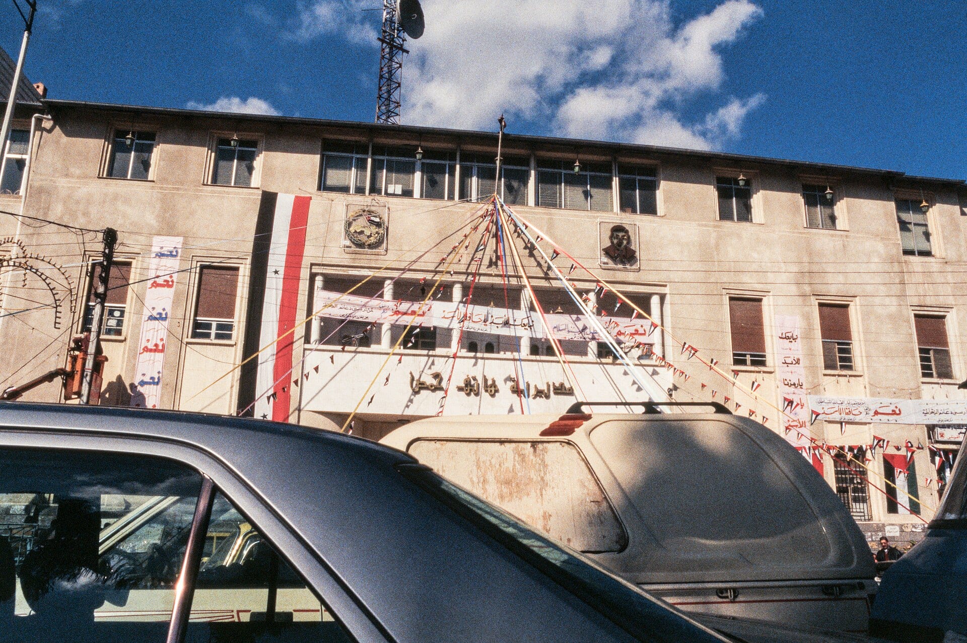 A street in Homs, traffic and shopfronts along both sides.